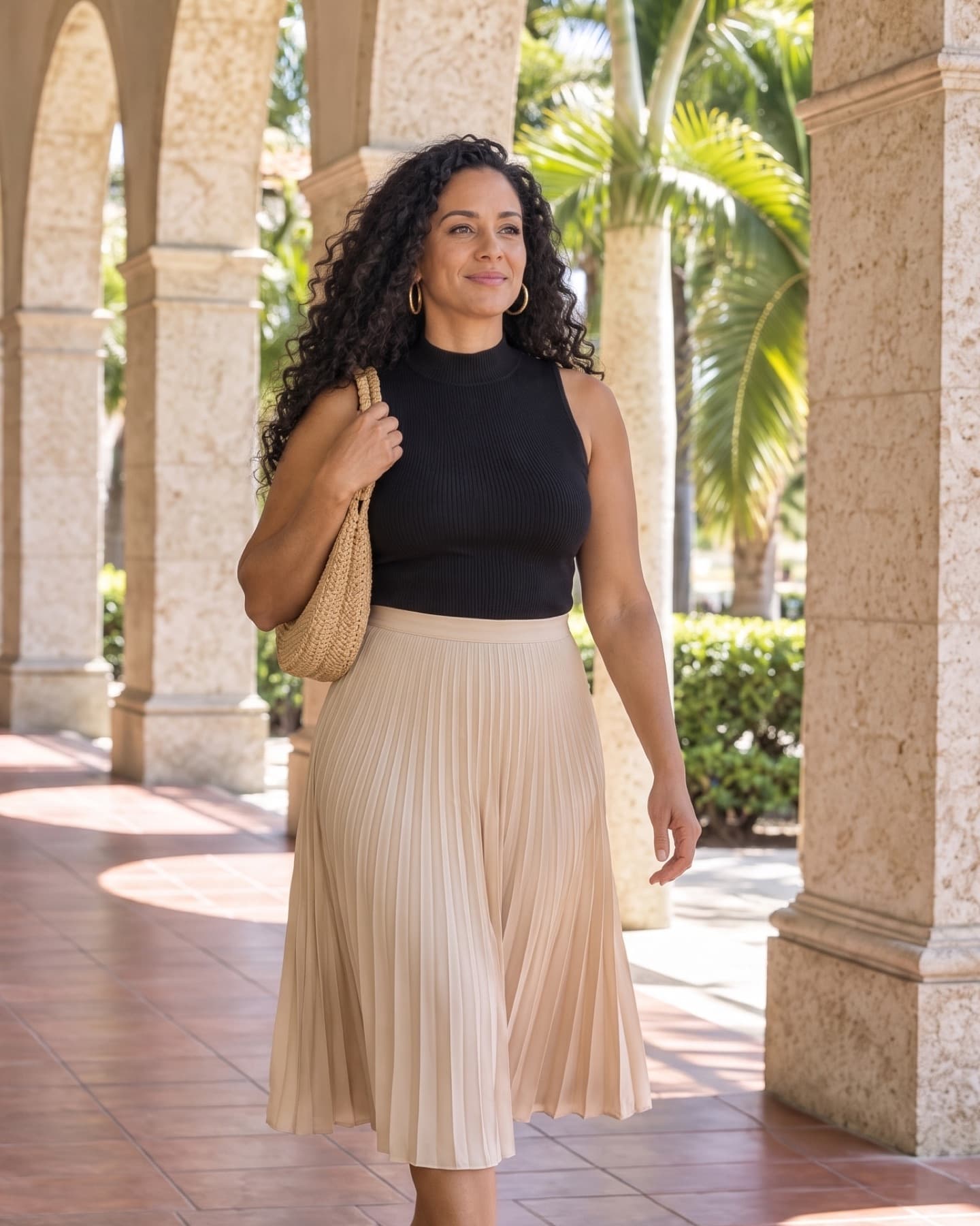 Woman with long dark curls in a black sleeveless high-neck top and a champagne pleated midi skirt, a woven bag on her shoulder, walking through a sunlit stone arcade with palms behind her